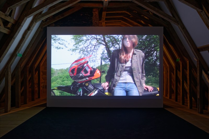 Laure Prouvost, The Smoking Image, 2015, tapisserie, vidéo (8.40 min), motos, machines à fumée, terre, moquette, œufs, smartphones, plumes.  Vue de l’exposition MDAC Rochechouart.  Courtesy de l’artiste, de MOT international (Londres, Bruxelles) et de la Galerie Nathalie Obadia (Paris, Bruxelles). Photo Aurélien Mole